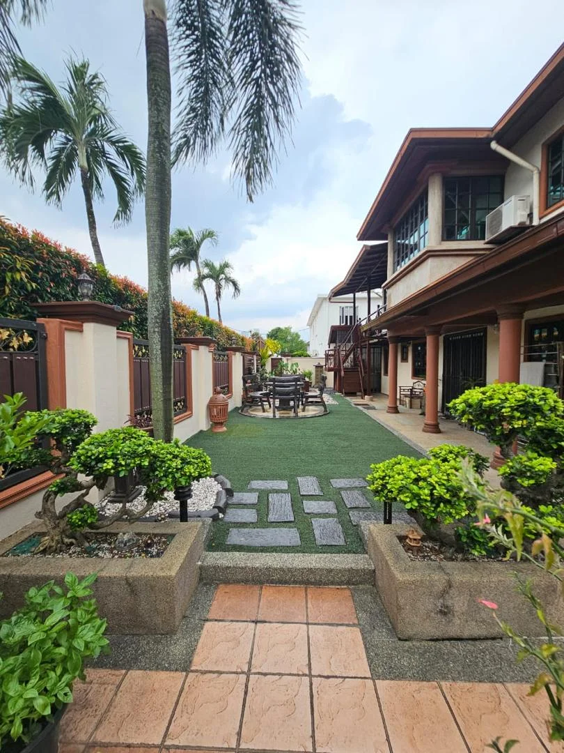 Exterior type: detached bungalow; features: manicured lawn, paved stone walkway, tropical landscaping, potted plants, outdoor dining area with table and chairs, covered patio, fence surrounding the property, palm trees, decorative stone planters; style: tropical-modern.